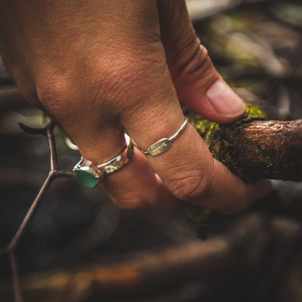 A woman's hand featuring two sterling silver rings. One is a hammered sea turquoise sea glass ring, and one is a delicate ring featuring an embossed twig branch design inspired by forests and nature. The hand is holding a moss covered branch.