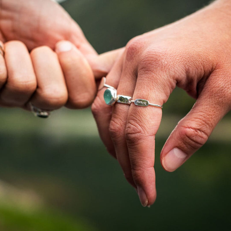 A woman's hand featuring two sterling silver rings. One is a hammered sea turquoise sea glass ring, and one is a delicate ring featuring an embossed twig branch design.