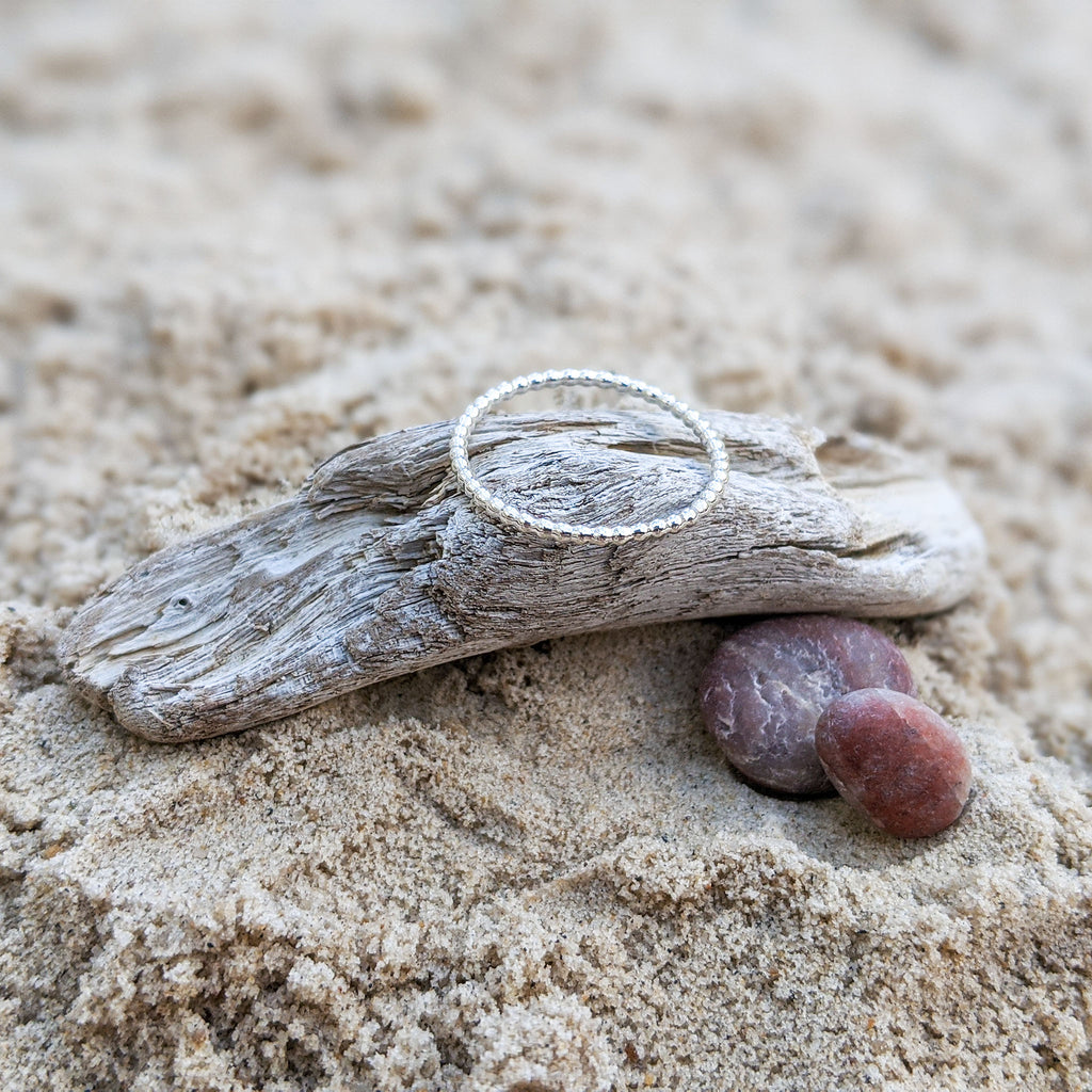 Sterling silver Beaded Droplet Ring resting on pale driftwood with pink stones and sand.