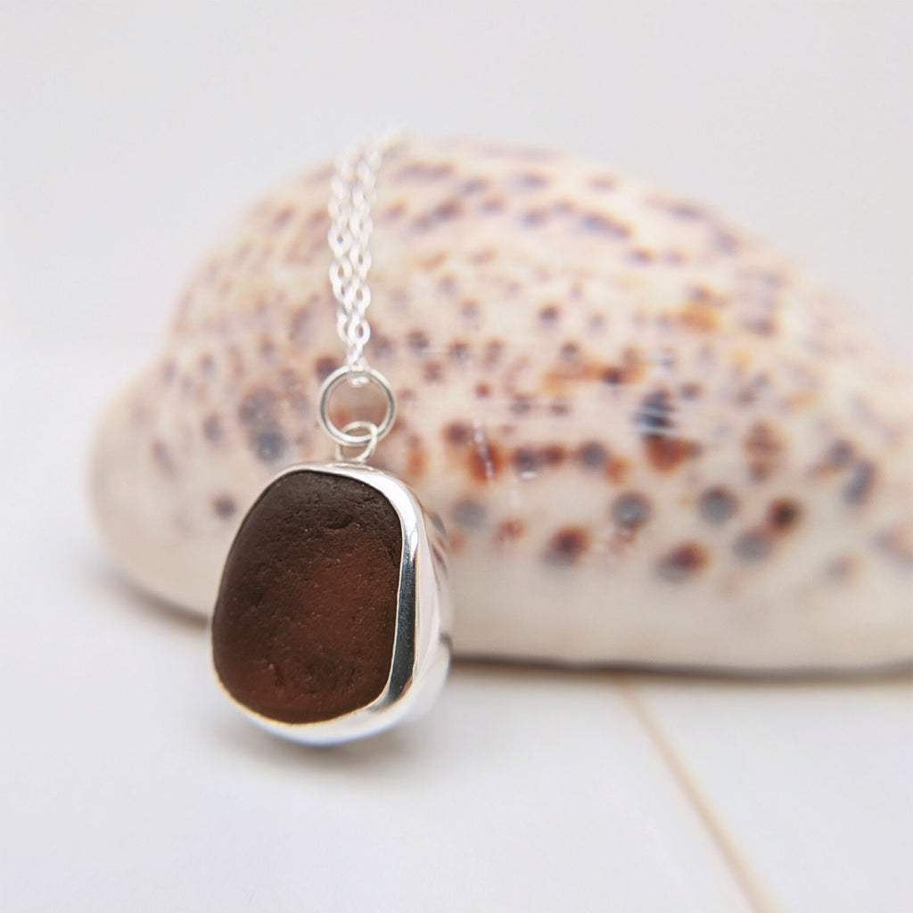 A close up of a brown earth-coloured sea glass necklace in a sterling silver bezel setting, resting on a shell