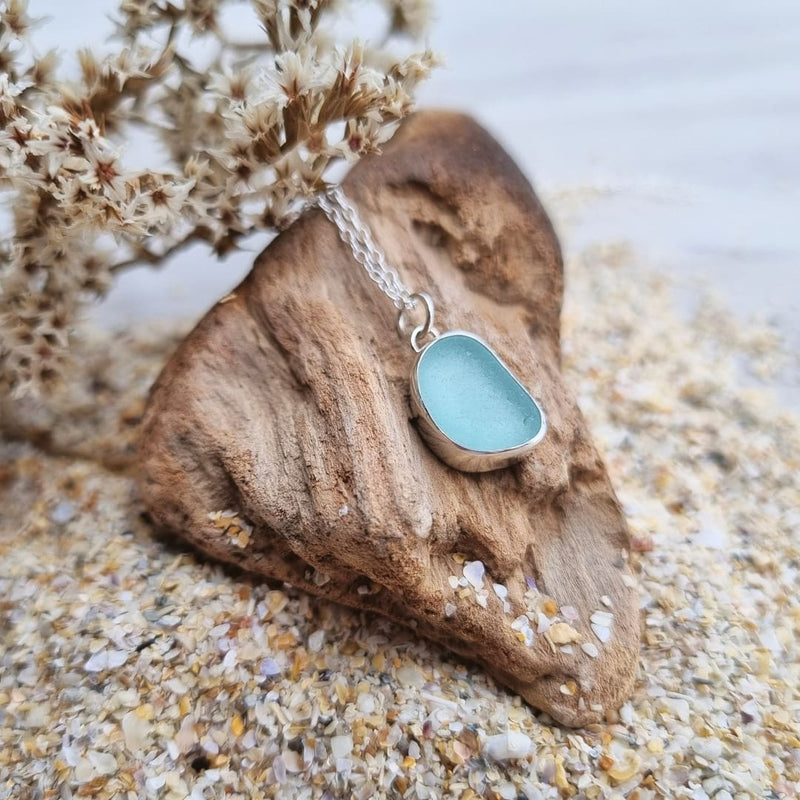 A close up photo of sterling silver sea glass necklace in a beautiful frosted blue hue, resting on a piece of driftwood and sand.