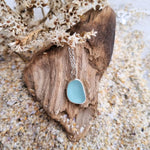 A close up photo of sterling silver sea glass necklace in a beautiful frosted blue hue, resting on a piece of driftwood and sand.