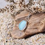 A close up photo of sterling silver sea glass necklace in a beautiful frosted blue hue, resting on a piece of driftwood and sand.