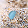 An extreme close up photo of sterling silver sea glass necklace in a beautiful frosted blue hue, resting on sand.