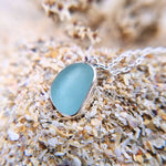 An extreme close up photo of sterling silver sea glass necklace in a beautiful frosted blue hue, resting on sand.