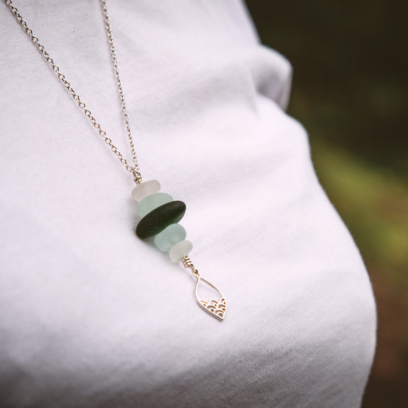 A close up photo of a sea glass stack necklace on a recycled sterling silver chain. The sea glass is white, forest green and sea foam colour, and the necklace is finished with a wave drop deco-style charm.