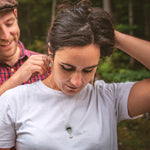 A photo of a man fastening a sea glass stack necklace around a woman's neck. The woman is looking down at the necklace. The necklace is on a recycled sterling silver chain. The sea glass is white, forest green and sea foam colour, and the necklace is finished with a wave drop deco-style charm.