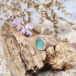 Photo of a turquoise sea glass ring with hammered sterling silver band, showcasing a vibrant turquoise hue reminiscent of ocean depths. The ring is resting on a piece of driftwood with a purple flower in the background.