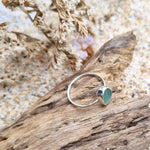 Photo from above of a turquoise sea glass ring with hammered sterling silver band, showcasing a vibrant turquoise hue reminiscent of ocean depths. The ring is resting on a piece of driftwood with coastal flowers in the background.