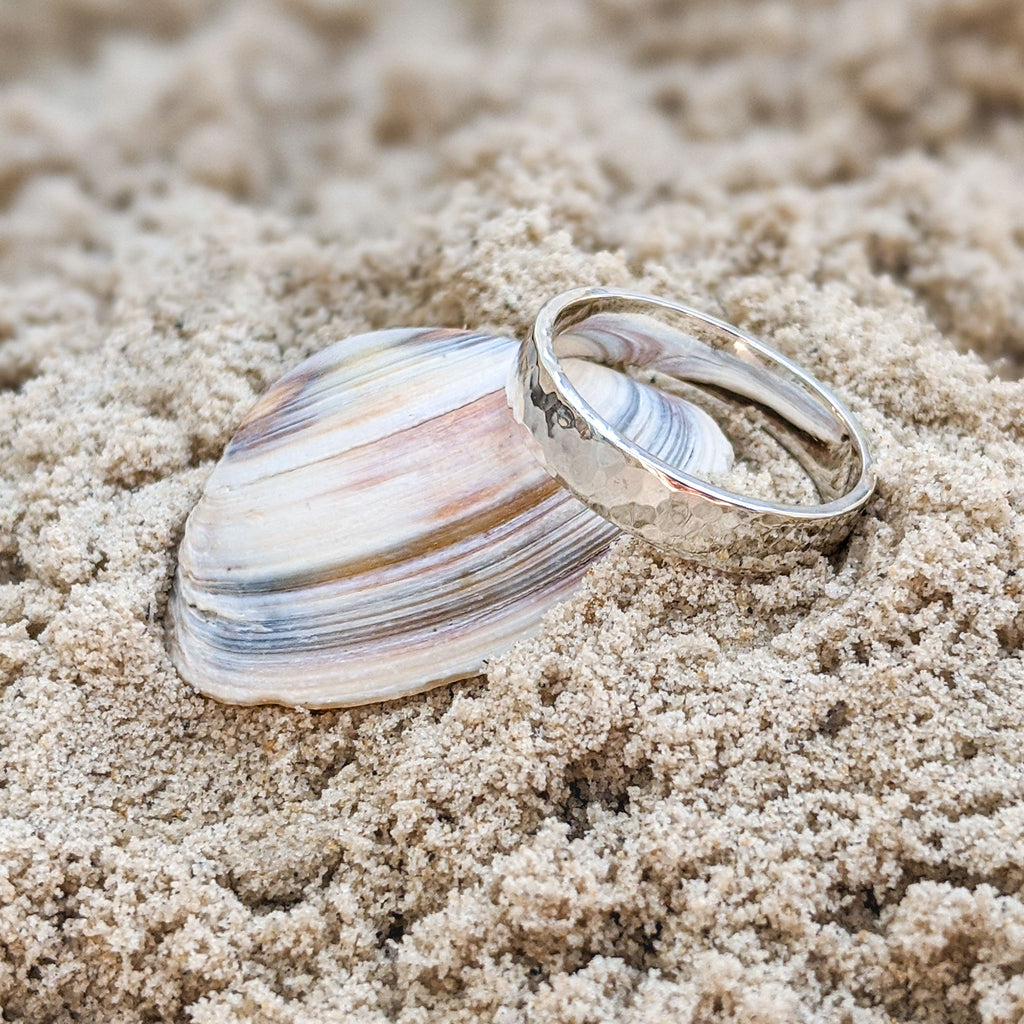 Close up of chunky sterling silver round ring with hammered texture design resting on pink and purple shell on sand 